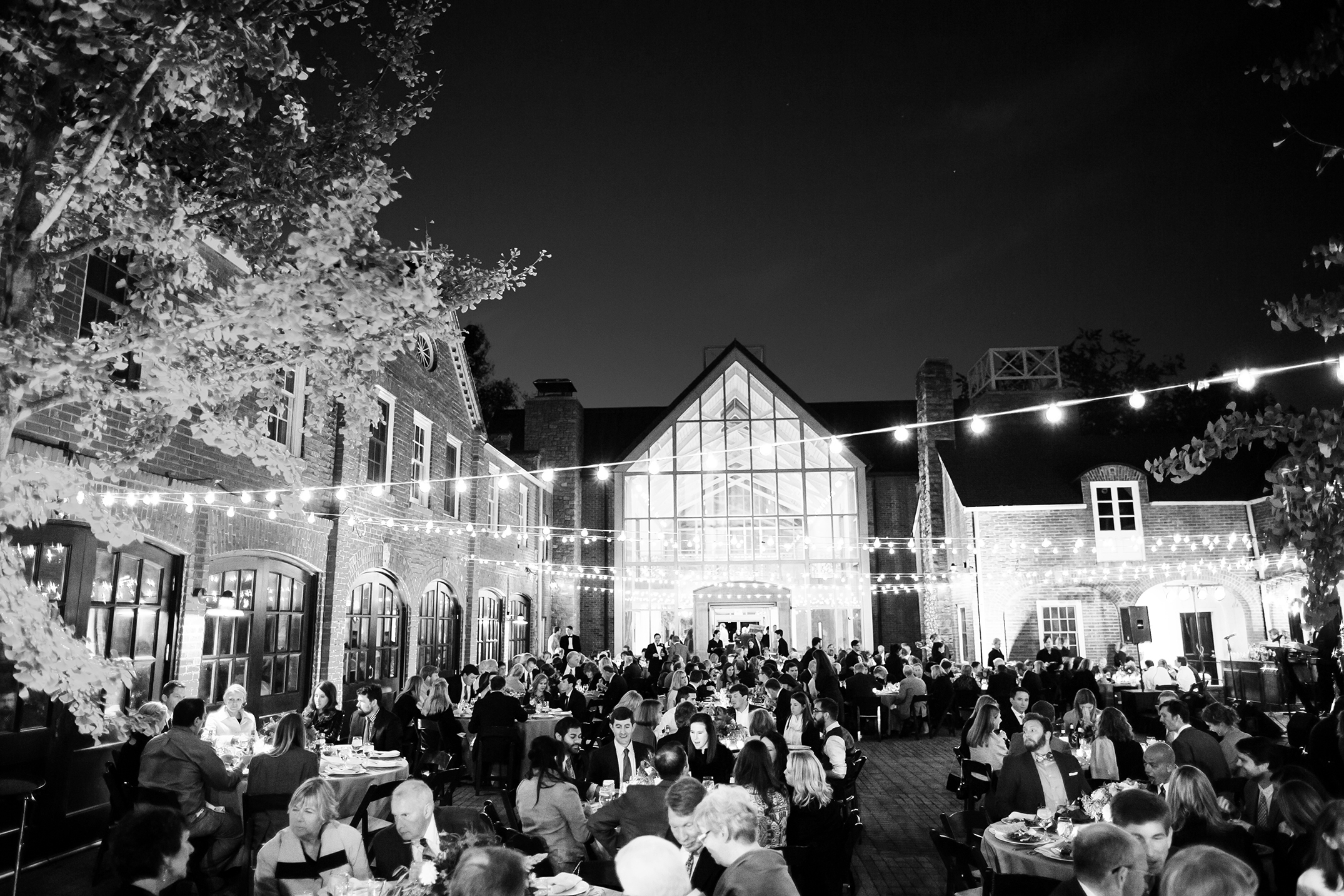 Cheekwood frist learning center at night with string lights and people seated for wedding reception. Image is in black and white.